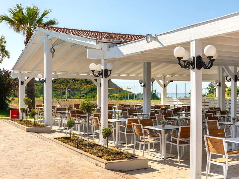 Gemütliche Terrasse mit weißem Dach, Holztischen und -stühlen, Palmen im Hintergrund.