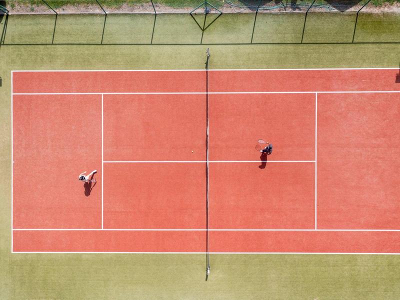 Draufsicht auf einen roten Tennisplatz mit zwei Spielern, umgeben von grünem Rasen.