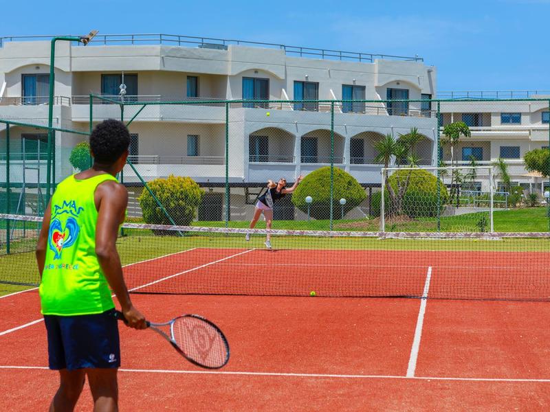 Zwei Personen spielen Tennis auf einem roten Sandplatz neben einem Hotel.