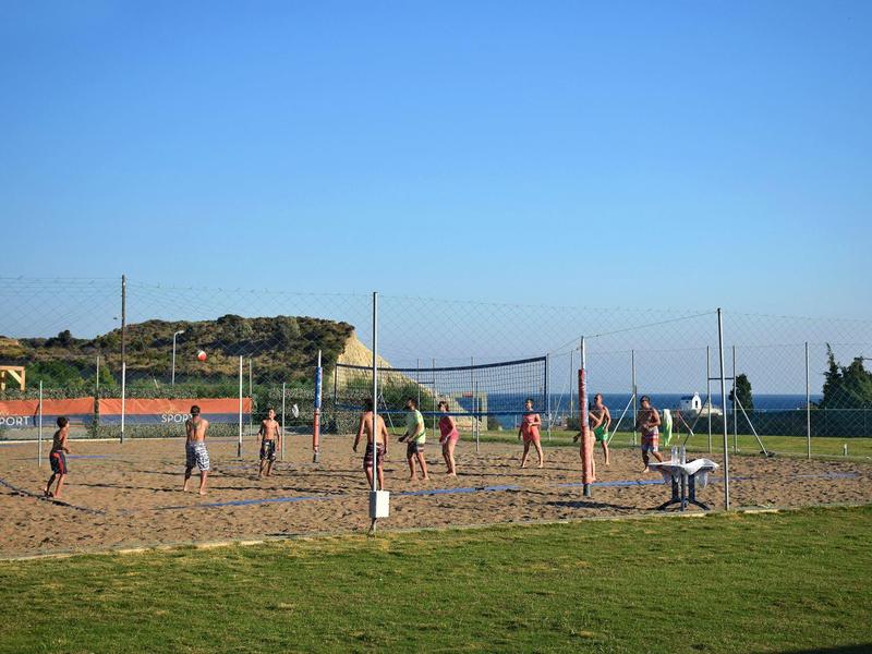 Menschen spielen Volleyball auf einem Sandplatz mit grünem Gras und klarem Himmel.