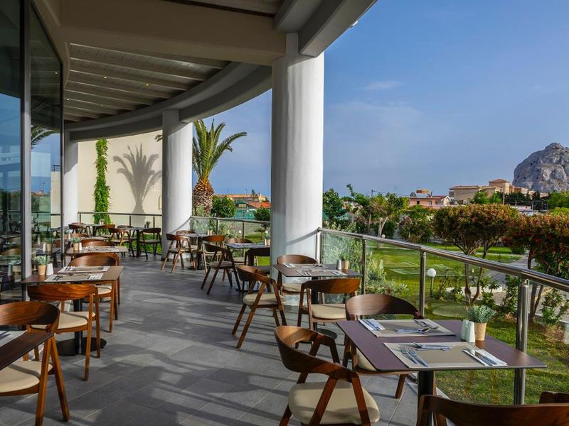 Terrasse mit Holzstühlen und Tischen neben Glasgeländer und Blick auf Garten und Berge.