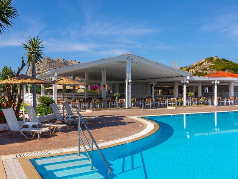 Poolbereich mit Liegestühlen vor einem Resort unter blauem Himmel.