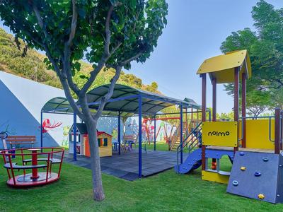 Kleurrijke speeltuin met klimrek, draaimolen en schommel omgeven door groene bomen.