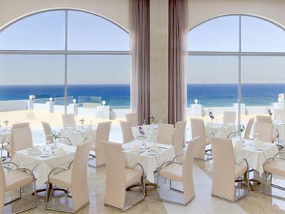 Restaurante elegante con sillas y mesas blancas con vista al mar a través de grandes ventanas.
