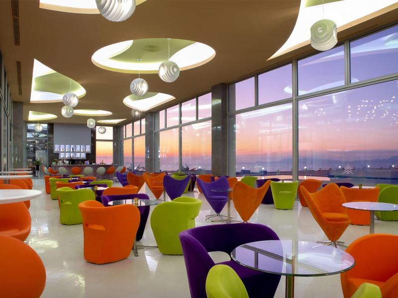 Kleurrijke lounge met moderne stoelen en grote ramen met uitzicht op zonsondergang.