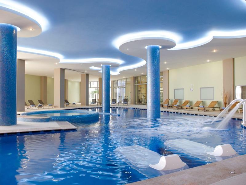 Overdekt zwembad met blauwe zuilen, watervallen en ligstoelen in een hotel.