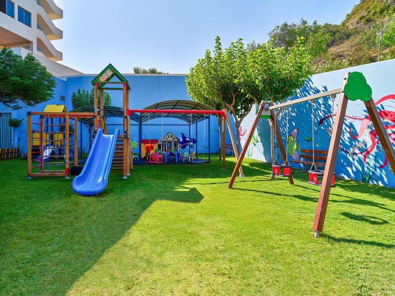 Speelplaats voor kinderen met glijbaan en schommel, omgeven door groen gras en een muur met kleurrijke muurschildering.