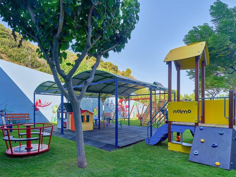 Kleurrijke speeltuin met klimrek, draaimolen en schommel omgeven door groene bomen.