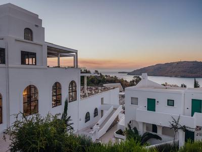 Weiße Gebäude mit grünen Fensterläden vor Meer und Hügeln bei Sonnenuntergang.