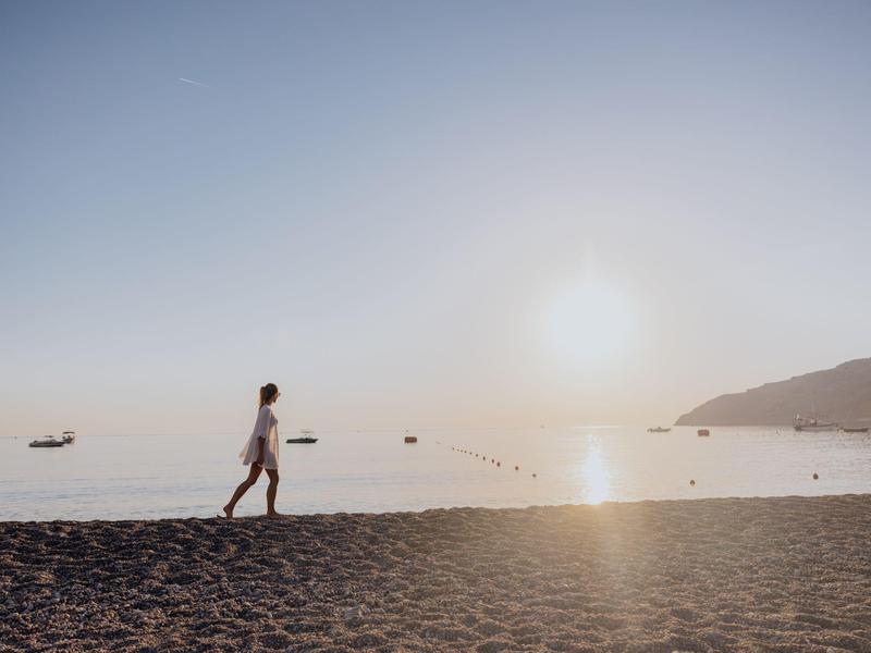 Strand mit Sand, ruhigem Meer, untergehender Sonne und person, die am Wasser entlang geht.