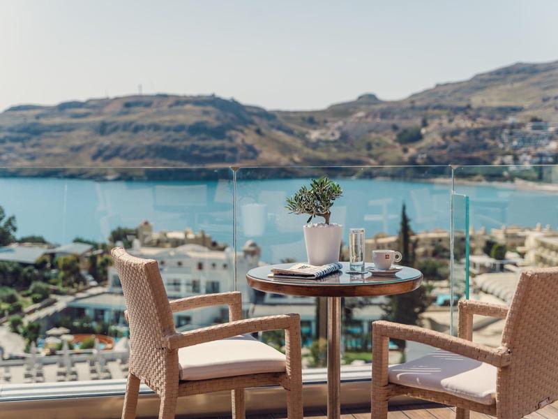 Balkon mit Holztisch und zwei Stühlen, Blick auf blaue Bucht und hügelige Landschaft im Hintergrund.