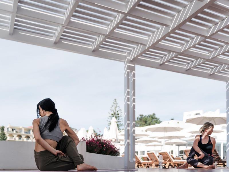 Zwei Frauen sitzen getrennt auf einer Terrasse mit Dach und weißen Möbeln, im Hintergrund Liegestühle.
