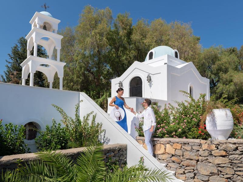 Weißes griechisches Kirchgebäude mit Glockenturm, Treppe und zwei Personen bei sonnigem Wetter.