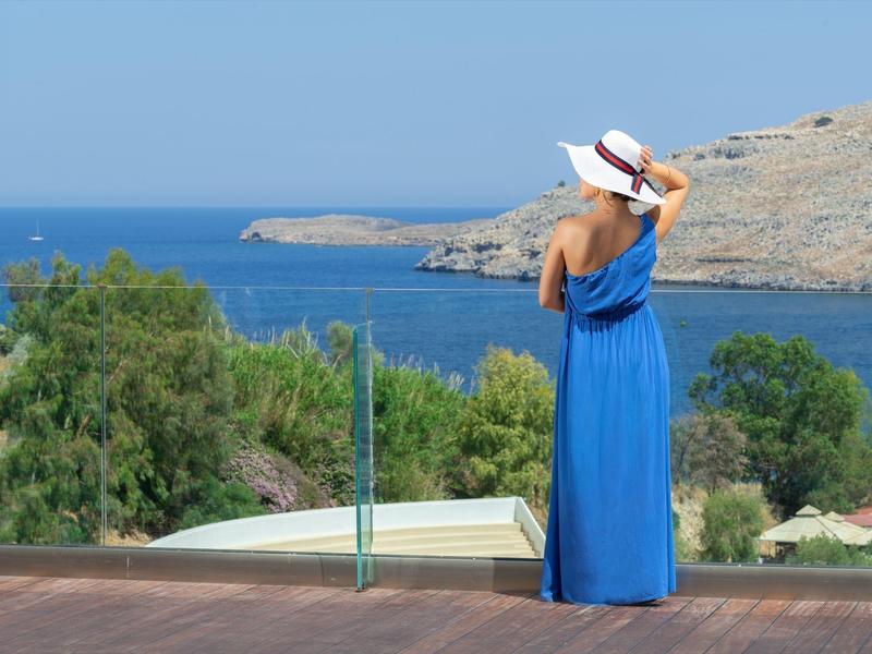 Frau in blauem Kleid mit weißem Hut blickt auf Meer und felsige Küste bei klarem Himmel.