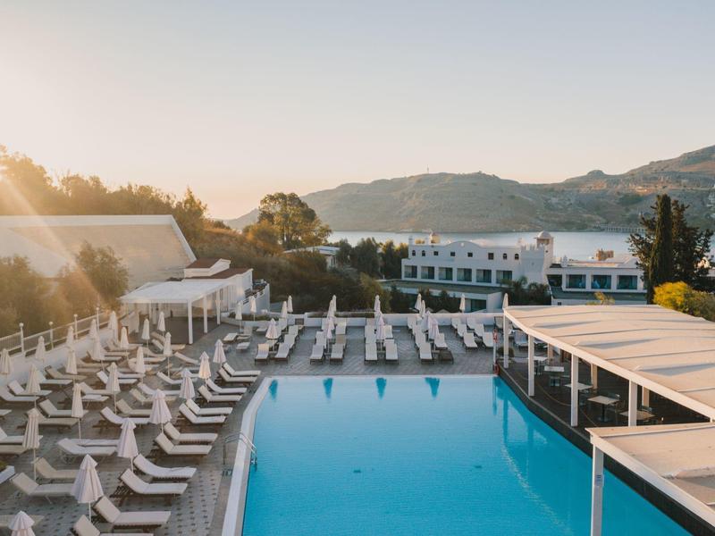 Großer Pool mit weißen Liegestühlen, Sonnenschirmen und umliegenden Gebäuden bei Sonnenuntergang.
