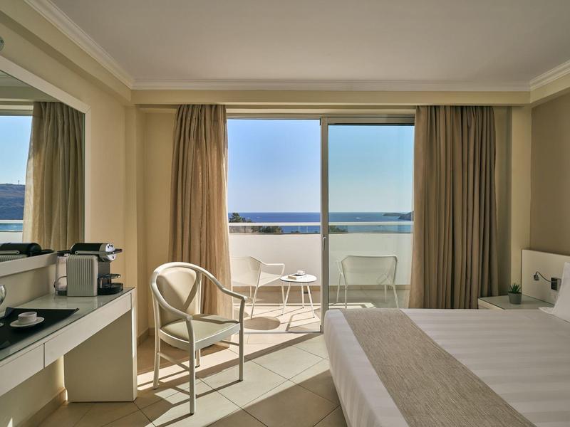 Helles Hotelzimmer mit Doppelbett, Stuhl, Balkon und Meerblick durch große Fenster.