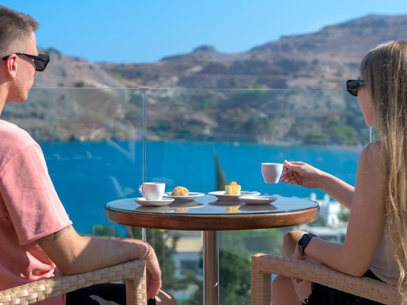 Zwei Personen sitzen am runden Tisch mit Kaffee vor blauem Wasser und Hügeln im Hintergrund.