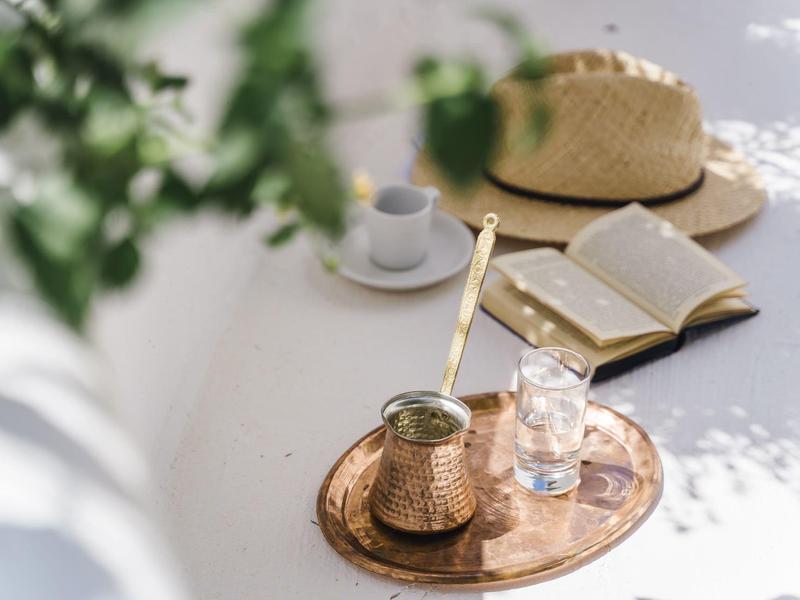Ein Tablett mit Kaffeekanne, Glas, offenes Buch und Hut auf einem weißen Tisch im Sonnenlicht.