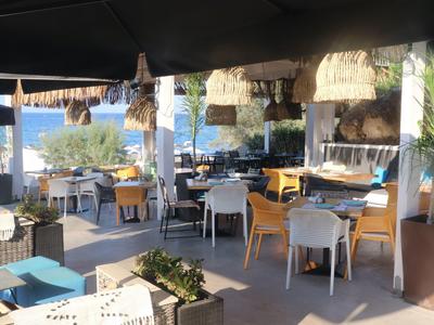 Offenes Café mit weiß-gelben Stühlen, Holztischen und Blick aufs Meer bei Sonnenschein.