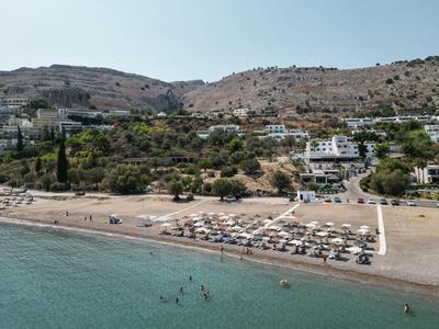 Strand mit Sonnenschirmen, ruhig blaues Meer, dahinter Hügel mit Vegetation und Gebäuden.