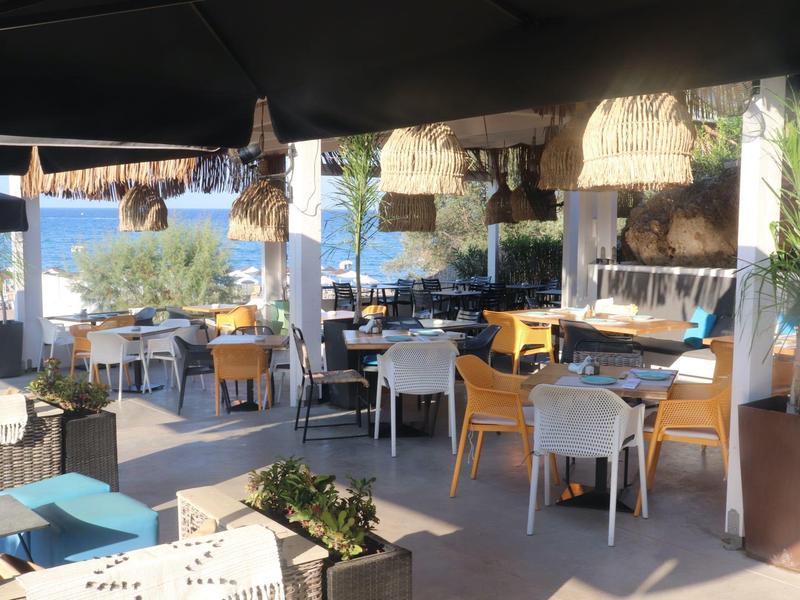Ristorante all'aperto con tavoli, sedie gialle e bianche, vista mare e ombrelloni.
