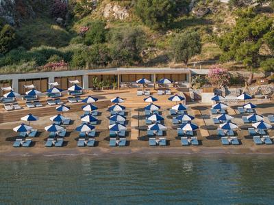 Strand mit Reihen von blauen Sonnenschirmen, Tischen und einem bewaldeten Hügel im Hintergrund.