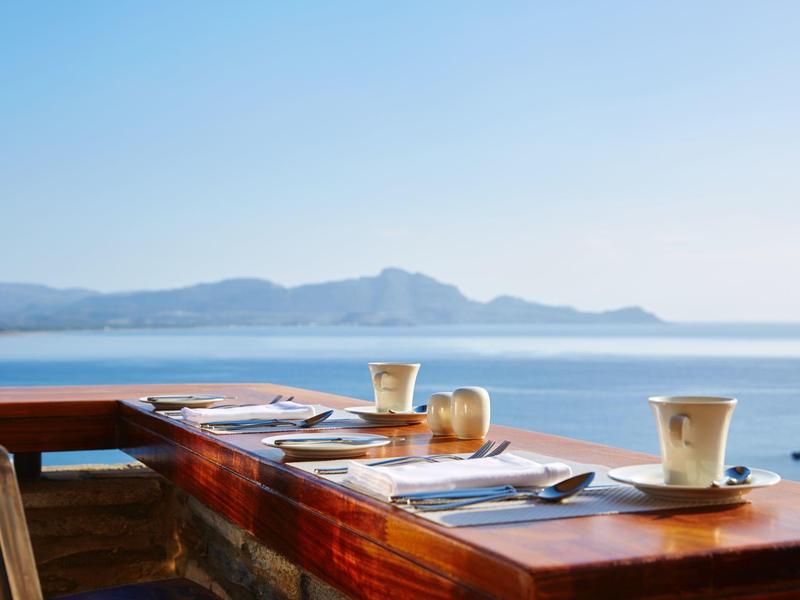 Ein Holzrestauranttisch mit Besteck und Tassen auf einer Terrasse mit Meerblick und Bergen im Hintergrund.