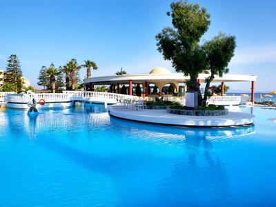 Großer blauer Pool mit Palmen und Menschen, umgeben von Gebäuden und sonnigem Himmel.