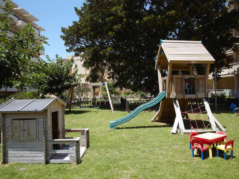 Kleiner Spielplatz mit Holzhaus, Schaukel, Rutsche und buntem Kindertisch auf grünem Rasen.