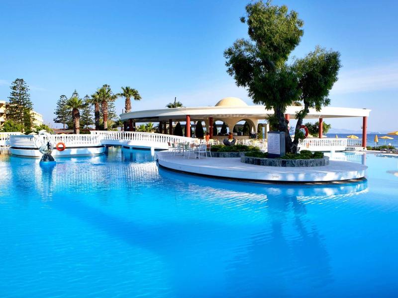 Großer blauer Pool mit Palmen und Menschen, umgeben von Gebäuden und sonnigem Himmel.