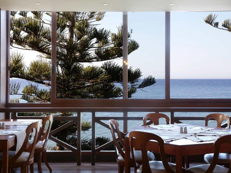 Restaurant mit Holztischen und Stühlen, Fenster mit Blick auf Meer und Nadelbaum im Hintergrund.