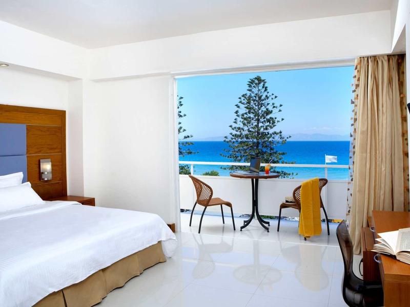Helles Hotelzimmer mit Doppelbett, Balkon mit Tisch und Stühlen, Blick aufs Meer und blauen Himmel.