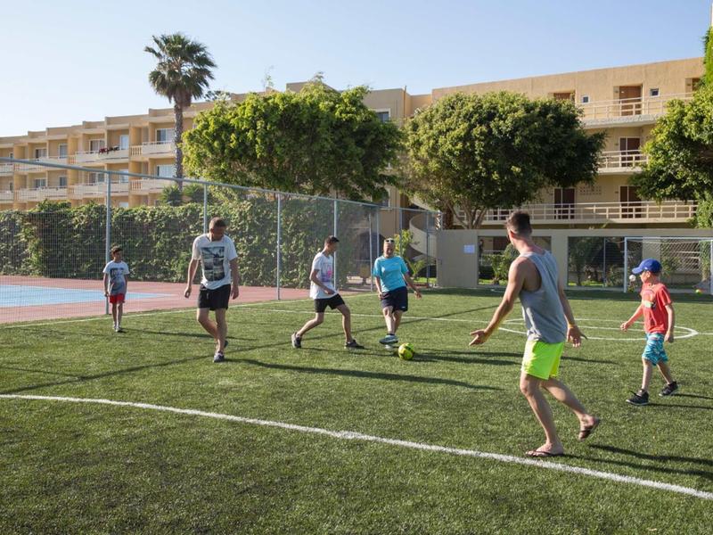 Mehrere Personen spielen Fußball auf einem grünen Spielfeld bei sonnigem Wetter vor einem Gebäude.