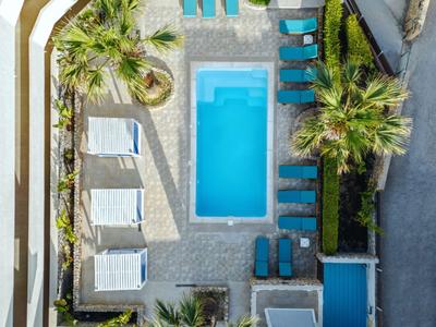 Vista aerea di un hotel con piscina blu, lettini e palme su pavimento grigio.