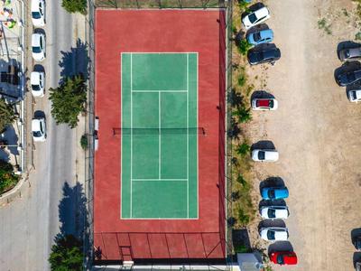 Un campo da tennis con superficie verde e bordo rosso, circondato da auto parcheggiate e una strada.