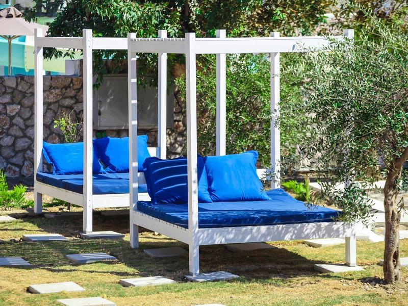 Zwei weiße Outdoor-Betten mit blauen Kissen in einem sonnigen Gartenbereich neben Steinmauer und Bäumen.