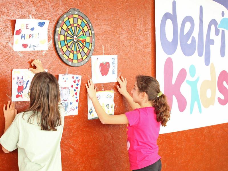 Zwei Mädchen hängen Kinderzeichnungen an eine Pinnwand neben einem Dartboard und einem Schild.
