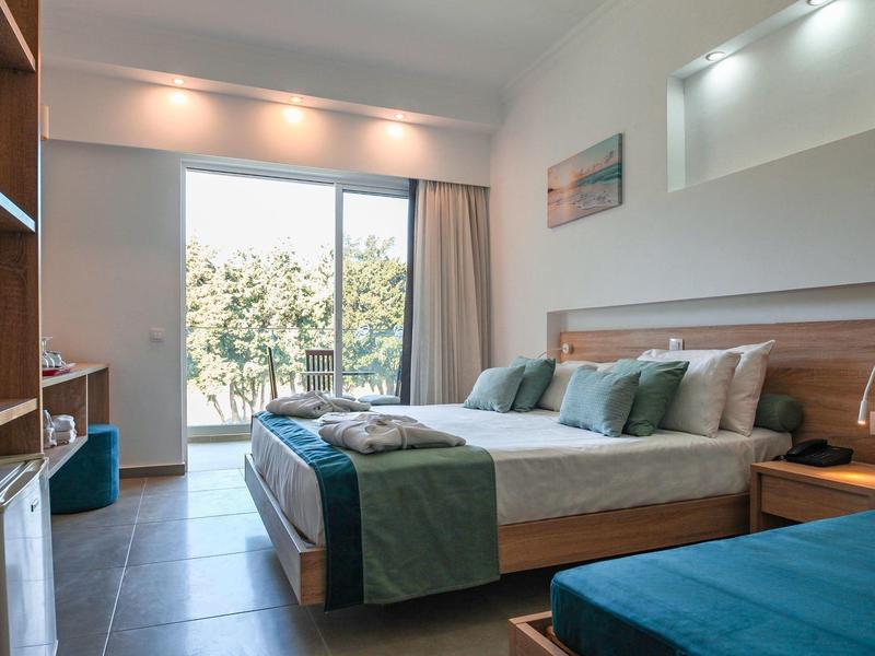 Helles Hotelzimmer mit Doppelbett, einem weiteren Bett, Holzmöbeln und Blick auf Bäume durch eine Glaszimmertür.