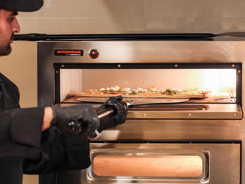 Mann schiebt Pizza in einen großen Edelstahlpizzaofen in einer Restaurantküche.