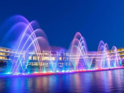 Beleuchtete Wasserfontänen vor einem modernen Hotel bei Nacht mit blauem Himmel.
