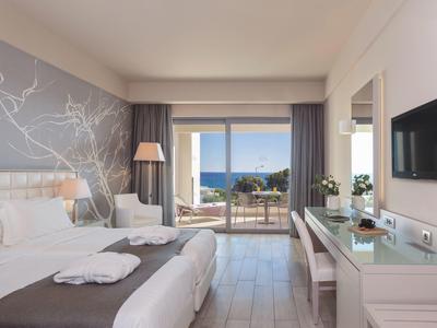Bright hotel room with double bed, desk, and sea view through large glass windows.