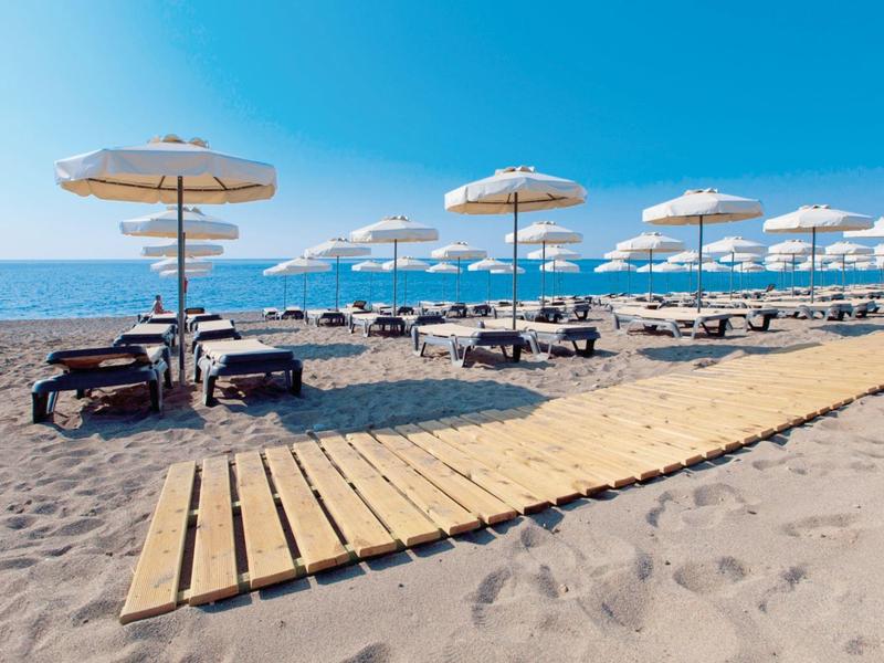 Sandy beach with sunbeds, umbrellas, and wooden walkway under a clear blue sky by the sea.
