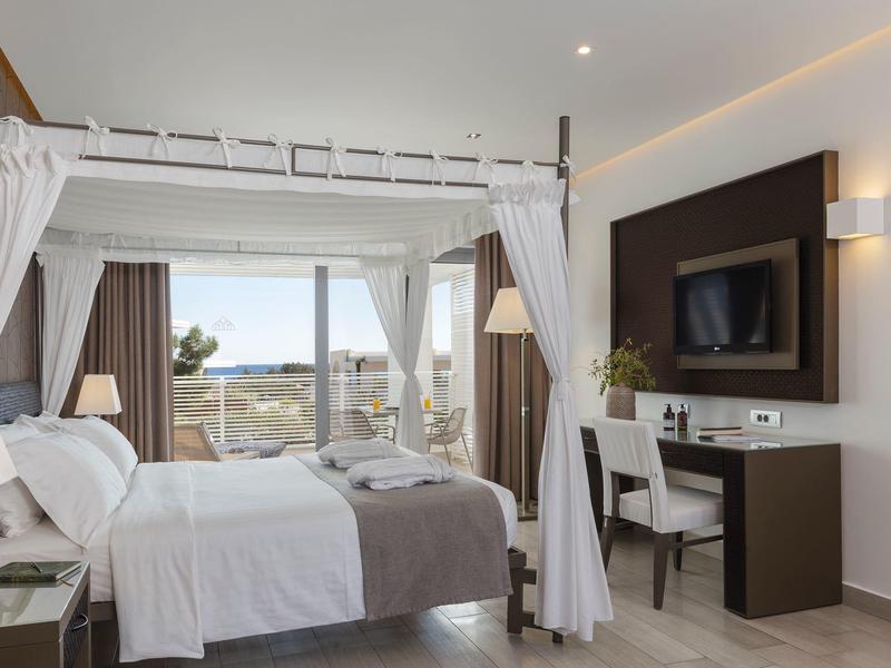 Bright hotel room with canopy bed, desk, and sea view through large windows.