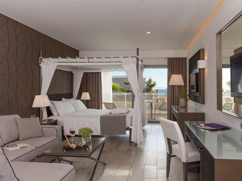 Modern hotel room with bed, sofa, TV, and balcony overlooking the sea.