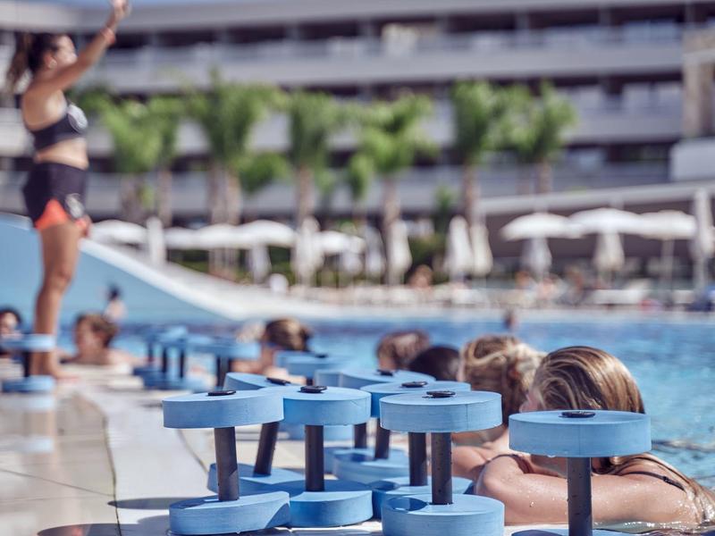 People in pool with blue water towers and hotel in background on sunny day.
