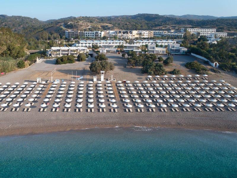 Large beach with many white sun loungers in front of a resort in a hilly area.