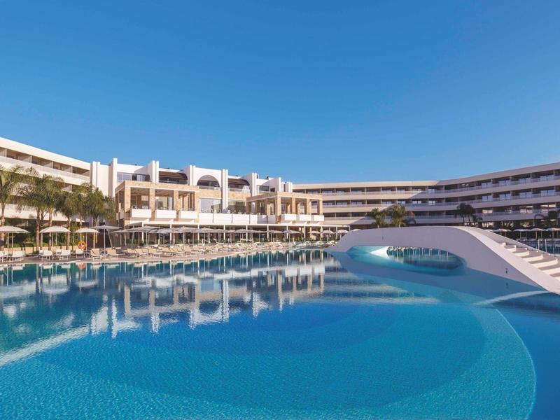 Large hotel pool with modern architecture and clear sky.