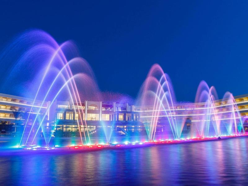 Colorful fountains in front of a modern hotel at night, reflected in the water.
