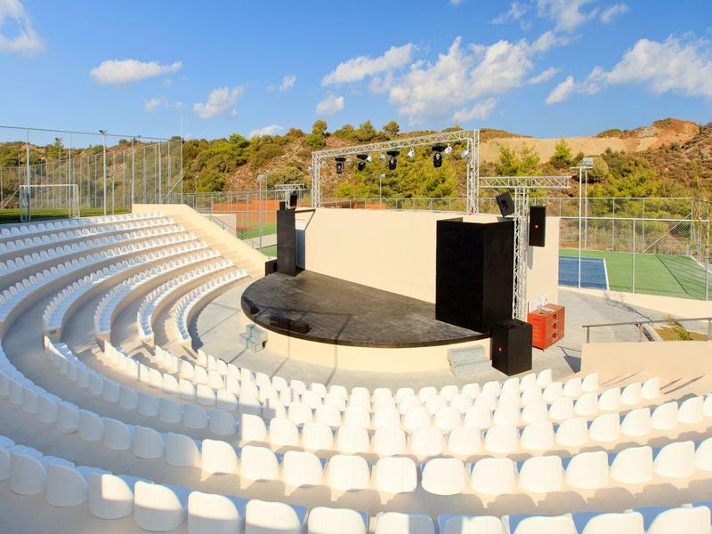 Open-air amphitheater with white seats and a stage against wooded hills.