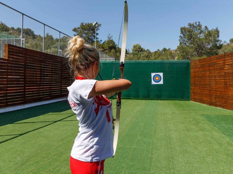 Woman practicing archery on green sports field aiming at a target on wooden wall.